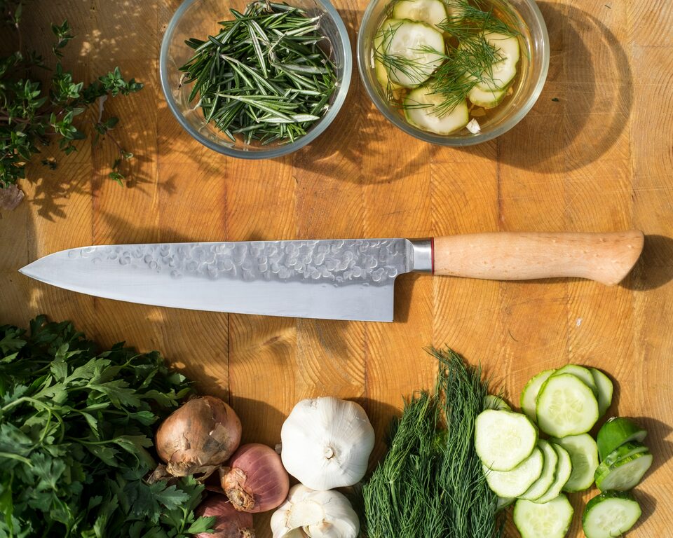 Kochmesser auf einem Holzbrett, daneben Kräuter, Zwiebeln, Knoblauch und Gurkenscheiben